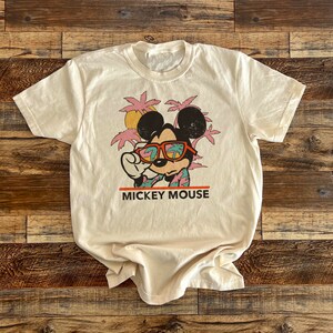 Puede incluir: Una camiseta beige con un gr&aacute;fico de estilo vintage de Mickey Mouse con gafas de sol y una camisa hawaiana. Tambi&eacute;n se incluyen palmeras y el texto "MICKEY MOUSE" en la camiseta.