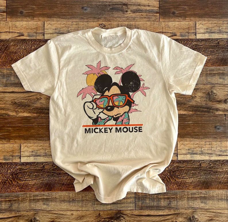 Puede incluir: Una camiseta beige con un gr&aacute;fico de estilo vintage de Mickey Mouse con gafas de sol y una camisa hawaiana. Tambi&eacute;n se incluyen palmeras y el texto "MICKEY MOUSE" en la camiseta.