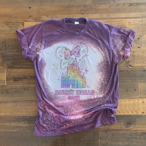 May include: Purple t-shirt with a bleach-splattered design featuring a cartoon drawing of Cinderella's castle with fireworks and a pink bow. The text "DISNEY WORLD 2023" is printed below the castle.