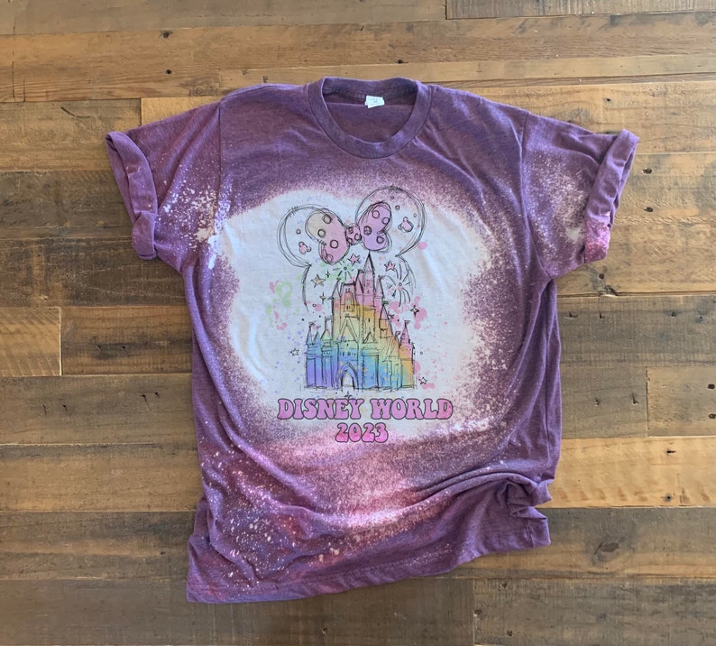 May include: Purple t-shirt with a bleach-splattered design featuring a cartoon drawing of Cinderella's castle with fireworks and a pink bow. The text "DISNEY WORLD 2023" is printed below the castle.