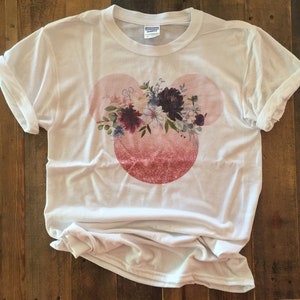 May include: White t-shirt with a pink and white floral design in the shape of Mickey Mouse ears.