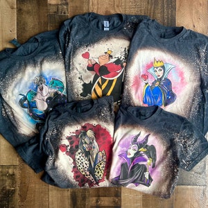 May include: Five dark gray t-shirts with bleach-splattered designs featuring Disney villains. The villains include Ursula, the Queen of Hearts, the Evil Queen, Maleficent, and Cruella de Vil.