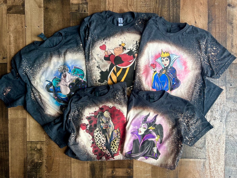 May include: Five dark gray t-shirts with bleach-splattered designs featuring Disney villains. The villains include Ursula, the Queen of Hearts, the Evil Queen, Maleficent, and Cruella de Vil.