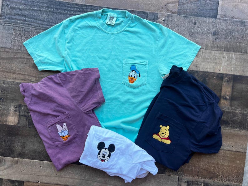 May include: Several t-shirts in shades of teal, purple, white, and navy blue. Each shirt has a pocket with an embroidered Disney character: Donald Duck, Daisy Duck, Mickey Mouse, and Winnie the Pooh. The shirts are laid out on a wooden surface.