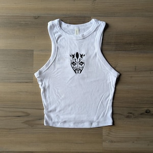 May include: A white sleeveless tank top featuring a black graphic print of a stylized face with intricate tribal-like markings centered on the chest. The garment has a ribbed crew neckline and wide armholes, constructed from a solid white fabric.