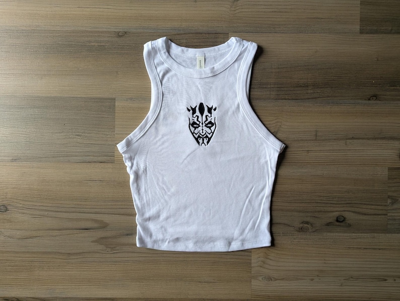 May include: A white sleeveless tank top featuring a black graphic print of a stylized face with intricate tribal-like markings centered on the chest. The garment has a ribbed crew neckline and wide armholes, constructed from a solid white fabric.