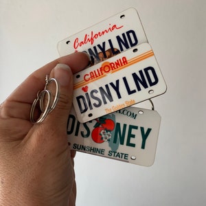 May include: Three white license plate-shaped keychains with the text "California" and "Disneyland" on them. The top keychain has the text "California" and "Disneyland" in black text. The middle keychain has the text "California" and "Disneyland" in red text. The bottom keychain has the text "Disney" in green text and "Sunshine State" in black text.