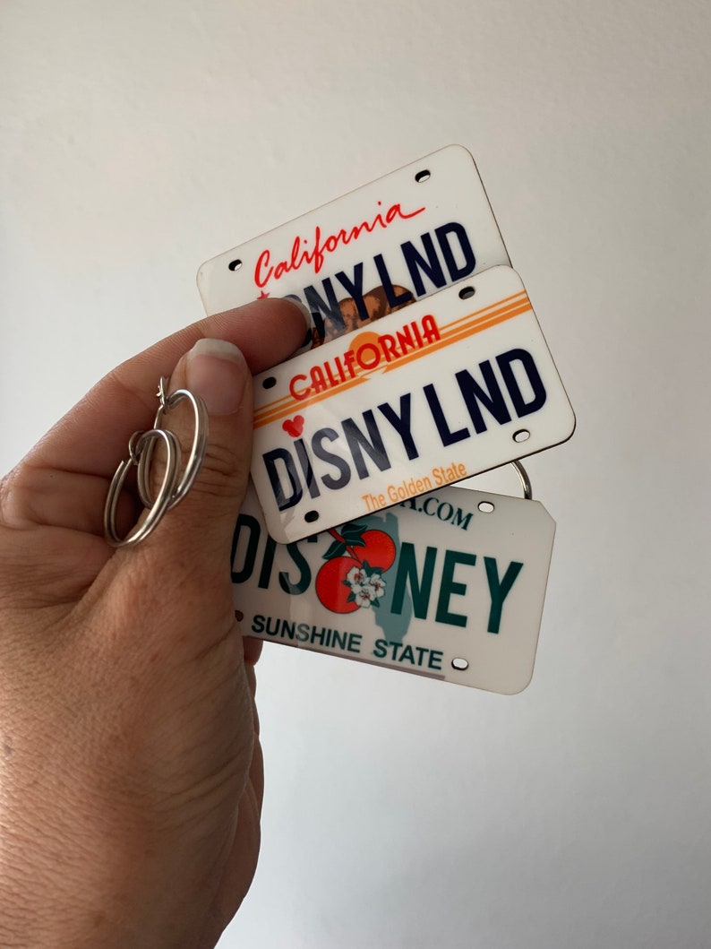 May include: Three white license plate-shaped keychains with the text "California" and "Disneyland" on them. The top keychain has the text "California" and "Disneyland" in black text. The middle keychain has the text "California" and "Disneyland" in red text. The bottom keychain has the text "Disney" in green text and "Sunshine State" in black text.