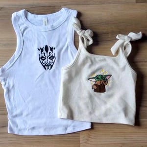 May include: A white sleeveless tank top featuring a black graphic of a horned character face and a cream-colored ribbed crop top with tie-shoulder straps, displaying an embroidered graphic of a small green creature in a brown robe with yellow stars.