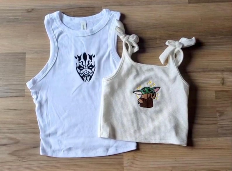 May include: A white sleeveless tank top featuring a black graphic of a horned character face and a cream-colored ribbed crop top with tie-shoulder straps, displaying an embroidered graphic of a small green creature in a brown robe with yellow stars.