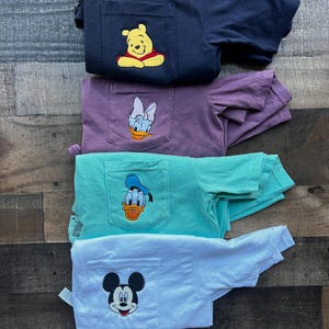 May include: Four folded t-shirts in navy, purple, teal, and white, each with a pocket featuring a cartoon character. The characters include Winnie the Pooh, Daisy Duck, Donald Duck, and Mickey Mouse. The shirts are stacked on a wooden surface.
