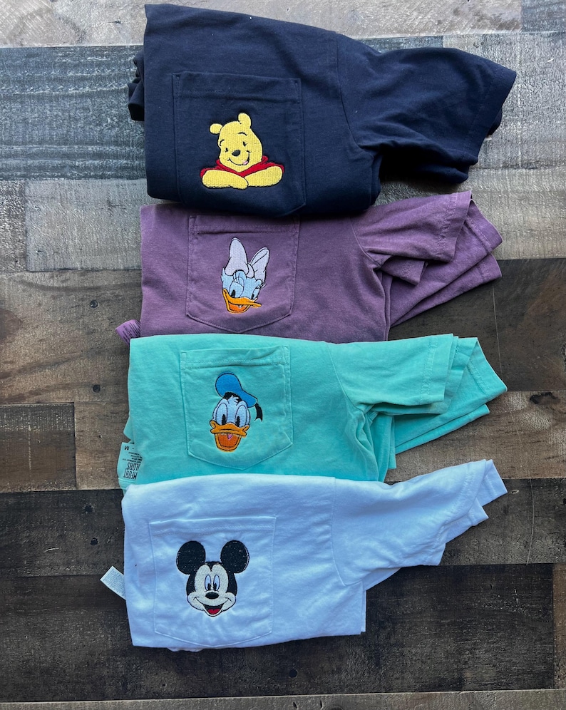 May include: Four folded t-shirts in navy, purple, teal, and white, each with a pocket featuring a cartoon character. The characters include Winnie the Pooh, Daisy Duck, Donald Duck, and Mickey Mouse. The shirts are stacked on a wooden surface.