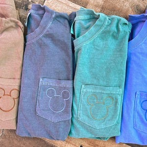 May include: Four folded t-shirts in shades of tan, blue, teal, and blue. Each shirt has a pocket with a Mickey Mouse head design. The shirts are made of a soft material and are likely for casual wear.