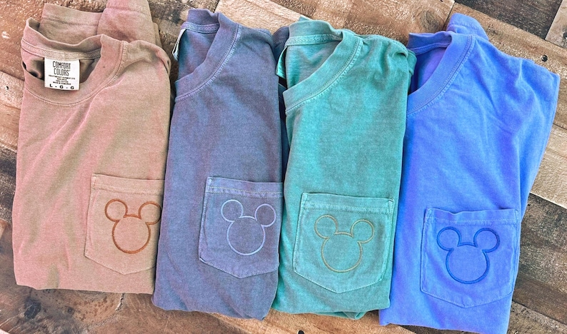 May include: Four folded t-shirts in shades of tan, blue, teal, and blue. Each shirt has a pocket with a Mickey Mouse head design. The shirts are made of a soft material and are likely for casual wear.
