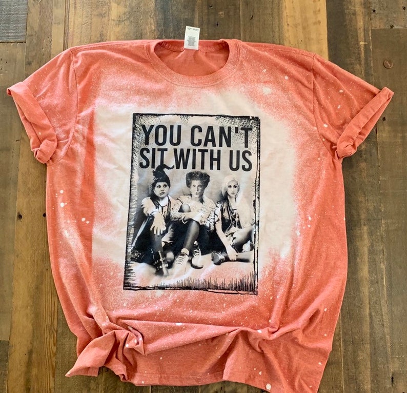 May include: A coral orange t-shirt with a bleached effect. The shirt features a black and white graphic of three women from the movie Hocus Pocus with the text "You Can't Sit With Us".