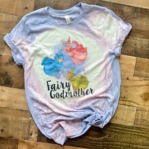 May include: A light blue t-shirt with a bleach-splattered design featuring three fairies flying with wands. The text "Fairy Godmother" is printed in black below the fairies.