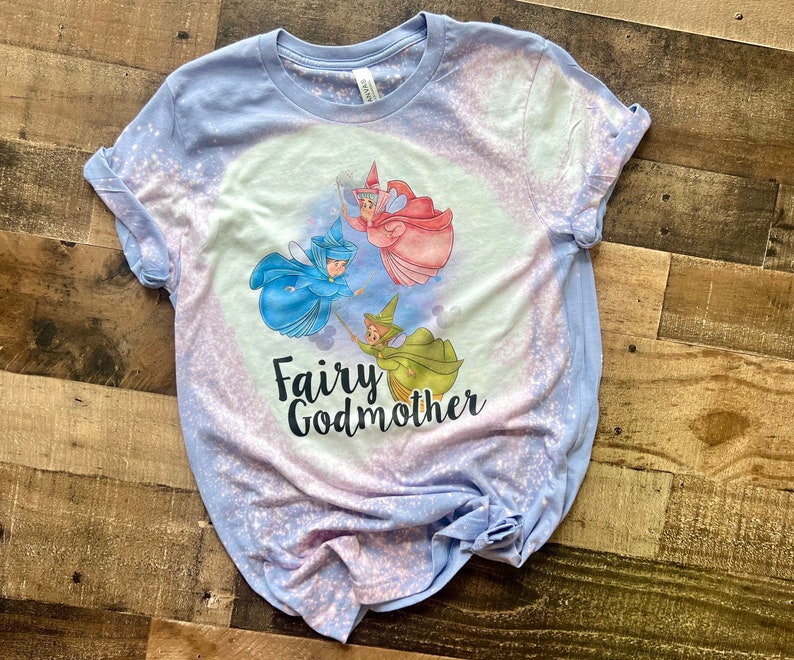 May include: A light blue t-shirt with a bleach-splattered design featuring three fairies flying with wands. The text "Fairy Godmother" is printed in black below the fairies.