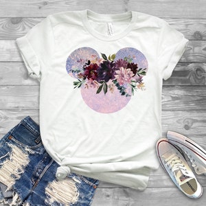 May include: White t-shirt with a floral design in shades of purple, pink, and green. The design is in the shape of Mickey Mouse ears.