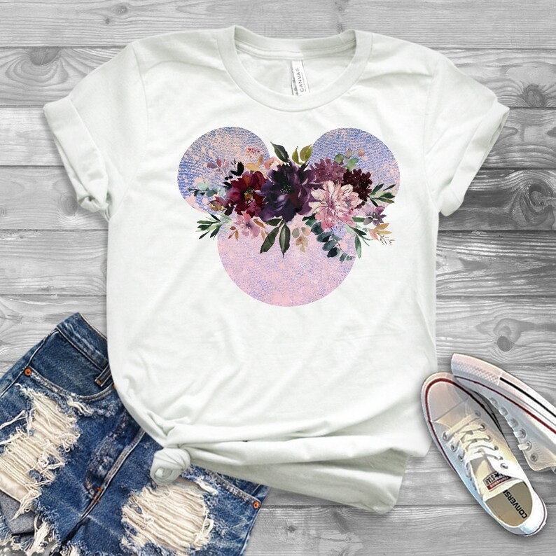 May include: White t-shirt with a floral design in shades of purple, pink, and green. The design is in the shape of Mickey Mouse ears.