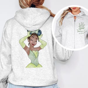 May include: A gray hooded sweatshirt with a zipper. The back of the sweatshirt features a cartoon illustration of Princess Tiana from Disney's The Princess and the Frog. The front of the sweatshirt has a small logo that says "Tiana & Co. est. 2007".