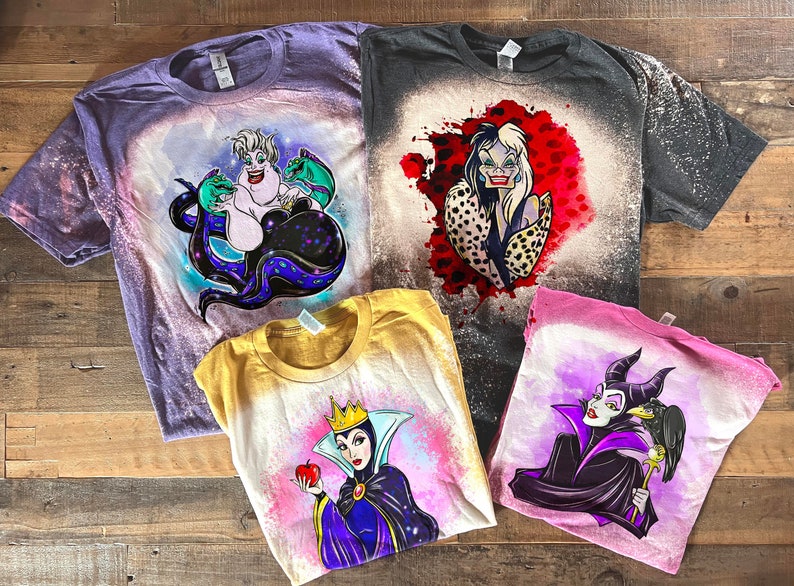 May include: Four bleach-dyed t-shirts featuring Disney villains. The shirts are purple, black, yellow, and pink. The villains are Ursula, Cruella de Vil, Evil Queen, and Maleficent.