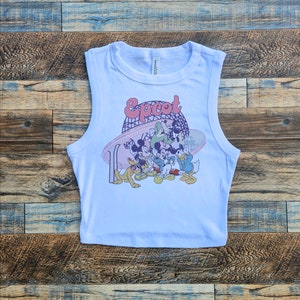 May include: White sleeveless crop top featuring a colorful graphic of Disney characters, including Mickey Mouse, Goofy, and Donald Duck, with the word "Epcot" in a retro font. The design includes a stylized depiction of a roller coaster.