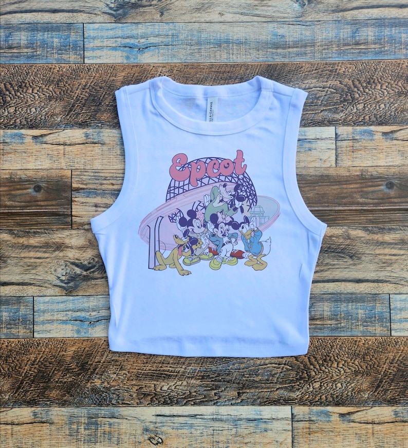 May include: White sleeveless crop top featuring a colorful graphic of Disney characters, including Mickey Mouse, Goofy, and Donald Duck, with the word "Epcot" in a retro font. The design includes a stylized depiction of a roller coaster.
