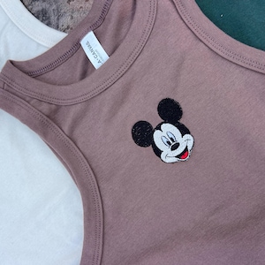 May include: Close-up of a mauve tank top with a Mickey Mouse embroidered design. The tank top has a round neckline and a sleeveless design. The tag reads "L.A. Canvas". Another white tank top is partially visible in the background.