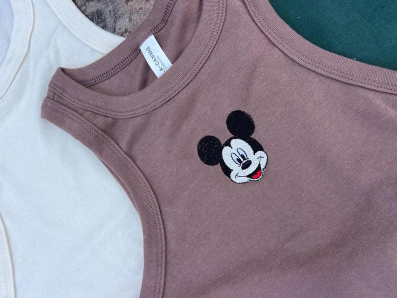 May include: Close-up of a mauve tank top with a Mickey Mouse embroidered design. The tank top has a round neckline and a sleeveless design. The tag reads "L.A. Canvas". Another white tank top is partially visible in the background.