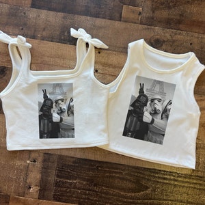 May include: Two white tank tops with a black and white photo print of Star Wars characters Darth Vader and a Stormtrooper in front of the Eiffel Tower. The photo shows the characters making a peace sign with their hands.