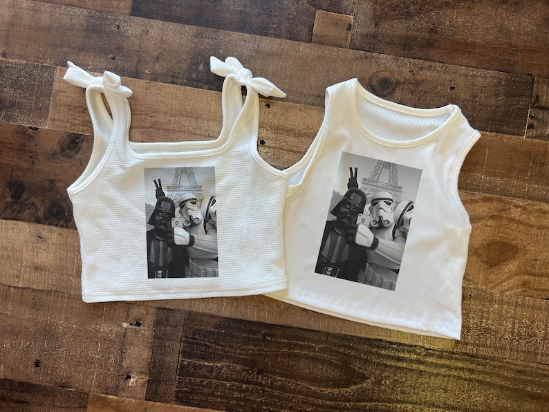 May include: Two white tank tops with a black and white photo print of Star Wars characters Darth Vader and a Stormtrooper in front of the Eiffel Tower. The photo shows the characters making a peace sign with their hands.