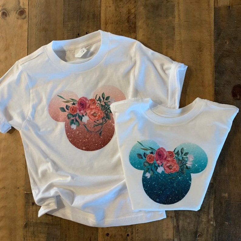May include: Two white t-shirts with a floral Mickey Mouse design. The larger shirt has a pink and orange glitter background, while the smaller shirt has a teal and blue glitter background.