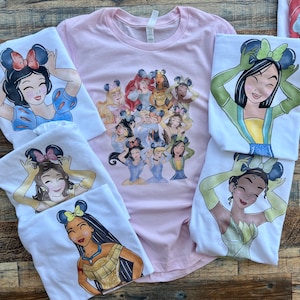 K&ouml;nnte beinhalten: Eine Kollektion von T-Shirts mit verschiedenen animierten Prinzessinnen, die Mausohr-Haarreifen tragen. Ein hellrosa T-Shirt zeigt eine Gruppe von Prinzessinnen, w&auml;hrend mehrere wei&szlig;e T-Shirts jeweils eine einzelne Prinzessin zeigen.