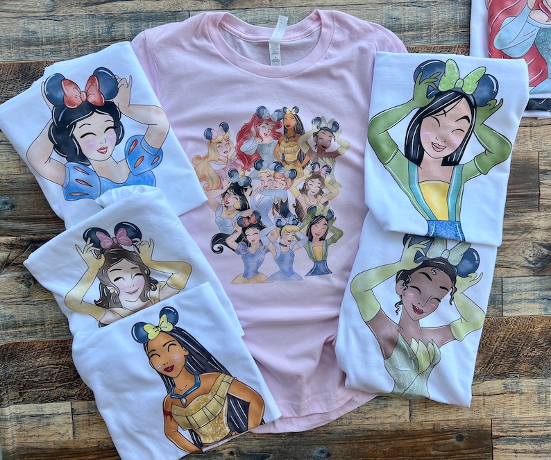 K&ouml;nnte beinhalten: Eine Kollektion von T-Shirts mit verschiedenen animierten Prinzessinnen, die Mausohr-Haarreifen tragen. Ein hellrosa T-Shirt zeigt eine Gruppe von Prinzessinnen, w&auml;hrend mehrere wei&szlig;e T-Shirts jeweils eine einzelne Prinzessin zeigen.