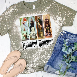 May include: A green t-shirt with a bleach-splattered design featuring a graphic of the Haunted Mansion characters from Disney. The text "Haunted Mansion" is printed in black below the graphic.