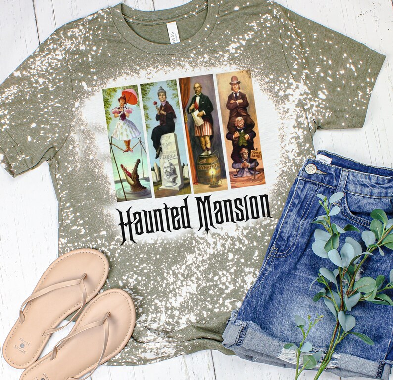 May include: A green t-shirt with a bleach-splattered design featuring a graphic of the Haunted Mansion characters from Disney. The text "Haunted Mansion" is printed in black below the graphic.
