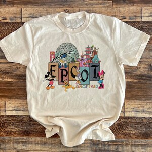 May include: A white t-shirt with a vintage-style graphic of Epcot, featuring Mickey Mouse, Donald Duck, Goofy, and other Disney characters. The graphic includes the text "Epcot Since 1982".