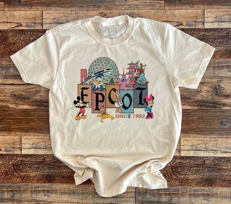 May include: A white t-shirt with a vintage-style graphic of Epcot, featuring Mickey Mouse, Donald Duck, Goofy, and other Disney characters. The graphic includes the text "Epcot Since 1982".