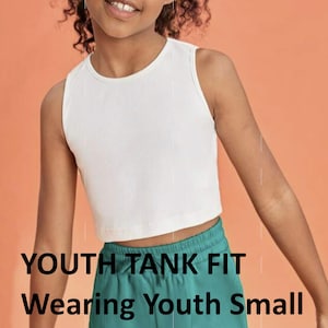 May include: White sleeveless tank top with a ribbed texture. The tank top is cropped and fits a youth size small. The model is wearing the tank top with green pants. The text "YOUTH TANK FIT" and "Wearing Youth Small" is visible at the bottom of the image.
