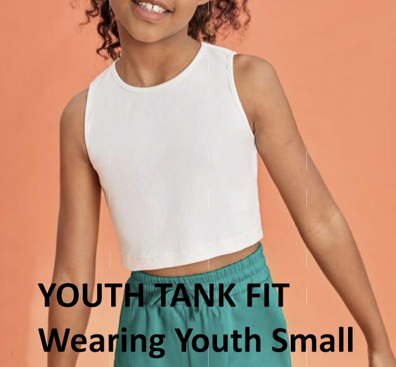 May include: White sleeveless tank top with a ribbed texture. The tank top is cropped and fits a youth size small. The model is wearing the tank top with green pants. The text "YOUTH TANK FIT" and "Wearing Youth Small" is visible at the bottom of the image.