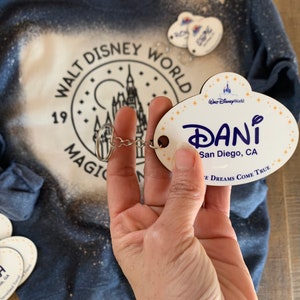 May include: White wooden name tag with the text "DANI" and "San Diego, CA" in blue lettering. The tag is attached to a silver key chain with a small silver charm. The tag has a blue and white Disney World logo in the top left corner and the text "Where Dreams Come True" in the bottom right corner.