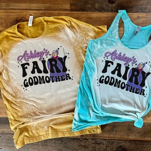 May include: Two bleached cotton shirts with a graphic design of a fairy godmother with the text "Ashley's Fairy Godmother". The shirt on the left is yellow and the shirt on the right is blue.