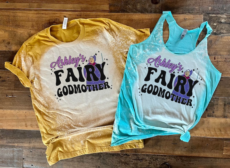 May include: Two bleached cotton shirts with a graphic design of a fairy godmother with the text "Ashley's Fairy Godmother". The shirt on the left is yellow and the shirt on the right is blue.
