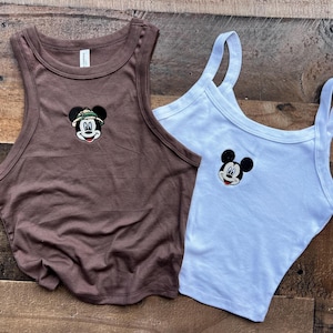 May include: A brown racerback tank top featuring an embroidered Mickey Mouse wearing a safari hat and a white tank top with an embroidered Mickey Mouse face. Both tops are sleeveless with a ribbed neckline and armhole trim, showcasing character art.