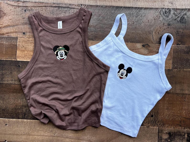 May include: A brown racerback tank top featuring an embroidered Mickey Mouse wearing a safari hat and a white tank top with an embroidered Mickey Mouse face. Both tops are sleeveless with a ribbed neckline and armhole trim, showcasing character art.