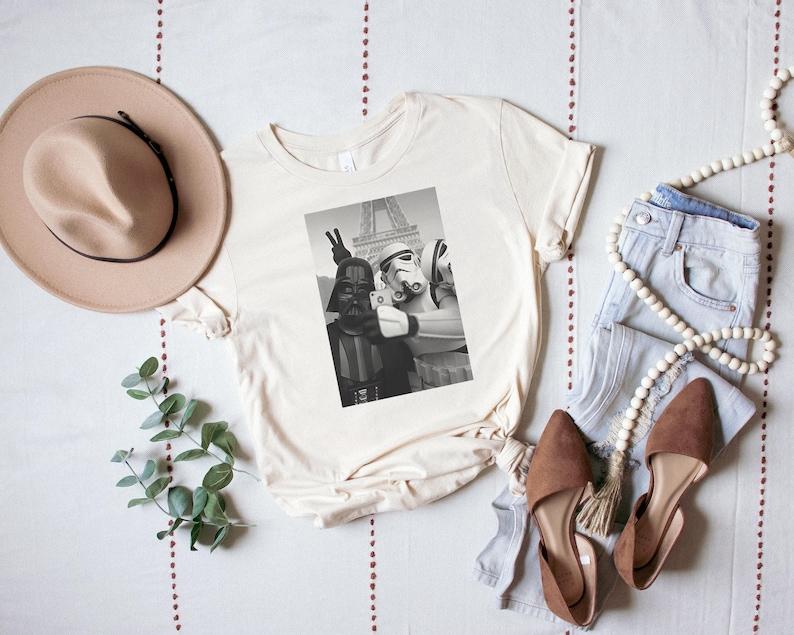 May include: A white t-shirt with a black and white photo print of Star Wars characters Darth Vader and Stormtroopers in front of the Eiffel Tower. The characters are making a peace sign and holding a camera.
