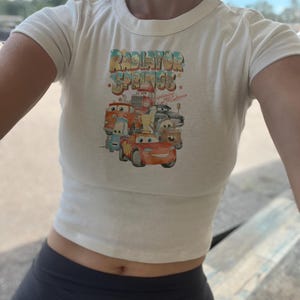 May include: White cropped t-shirt with a graphic of cartoon cars from the movie Cars. The graphic says "Radiator Springs" with the text "Home of the McQueen's" underneath.