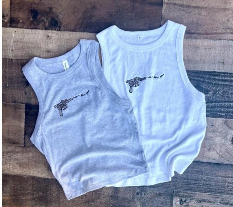 May include: A light gray tank top and a white tank top, both featuring a small black embroidered graphic of a ray gun firing a projectile with the text "pew pew" trailing from the barrel. Both garments have a ribbed crew neckline and sleeveless cut.
