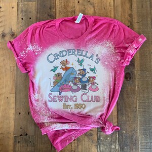 May include: Pink bleached t-shirt with a graphic of mice from Cinderella sewing. The text reads "Cinderella's Sewing Club Est. 1950".