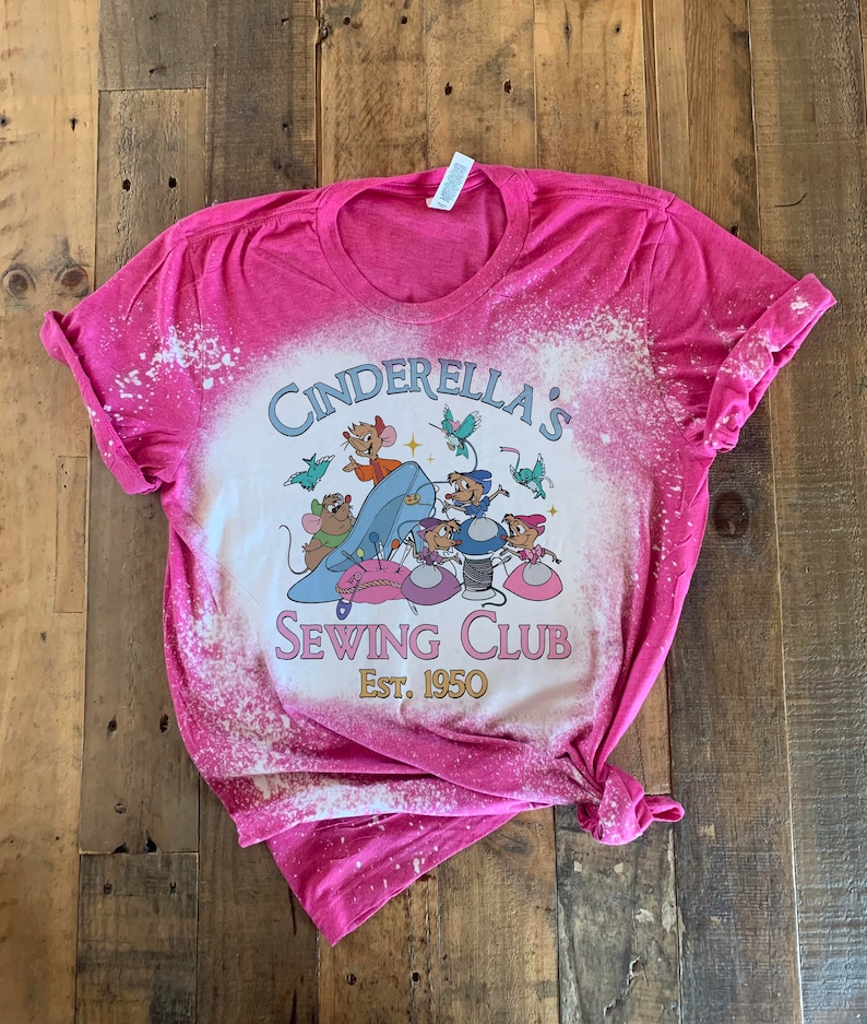 May include: Pink bleached t-shirt with a graphic of mice from Cinderella sewing. The text reads "Cinderella's Sewing Club Est. 1950".